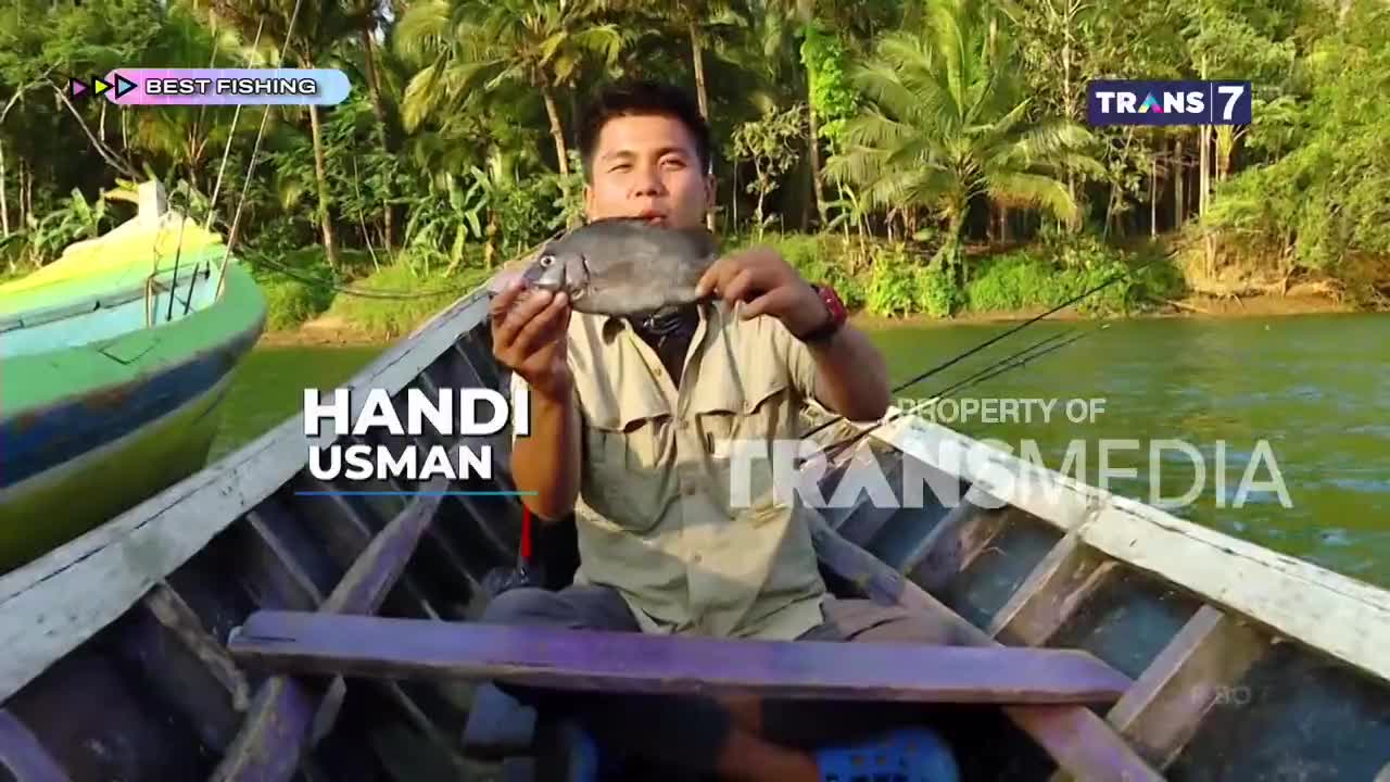 Handi Usman holds up a freshly caught fish from his boat on the river. The Trans7 crew captures the moment as he displays his catch against a backdrop of lush Indonesian greenery.