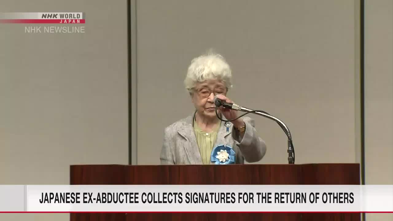 A woman with white hair, wearing a light-colored jacket, speaks into a microphone from behind a podium. The NHK World-Japan news graphic is visible in the corner, and the headline below reads "Japanese ex-abductee collects signatures for the return of others."
