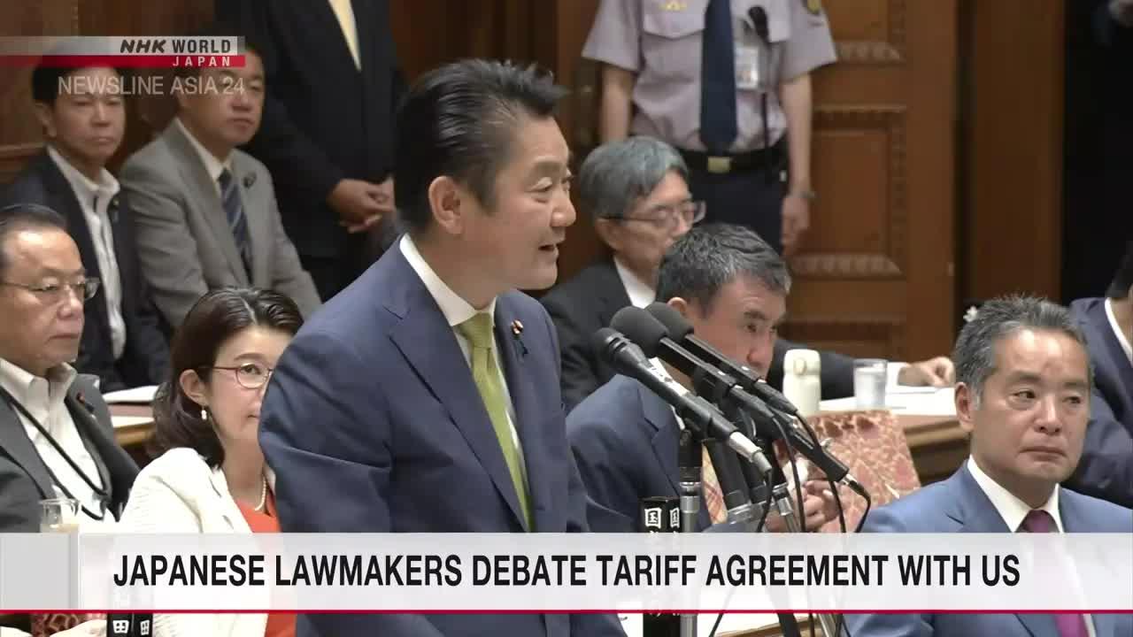 A man in a blue suit, speaking into multiple microphones, gestures with his hands. The NHK World-Japan news banner at the bottom announces the topic of the debate.
