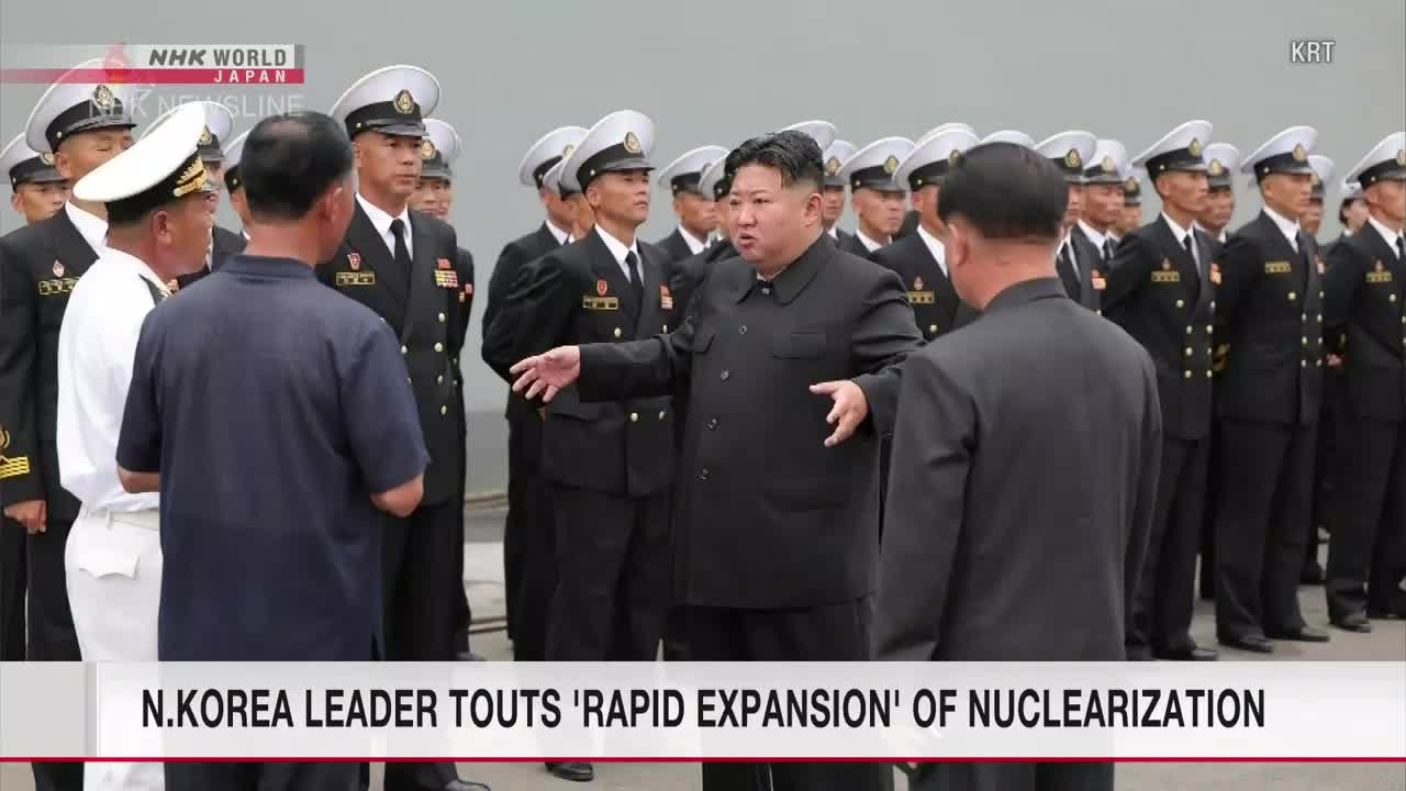 Kim Jong-un gestures emphatically, speaking to a group of men in dark suits and navy uniforms. Behind them, more uniformed personnel stand in neat rows, their white-topped hats catching the light.
