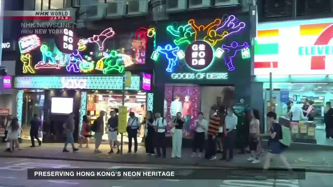 Neon signs, some in the shape of figures, illuminate the storefront of "Goods of Desire" as people mill about on the sidewalk. A news report from NHK World-Japan is covering the preservation of Hong Kong's neon heritage.
