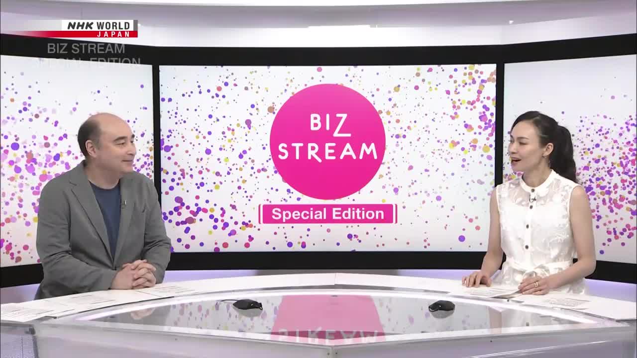 The two presenters on the NHK World-Japan program, *Biz Stream Special Edition*, are seated at a desk, facing each other. A large screen behind them displays the show's logo against a backdrop of colorful dots.
