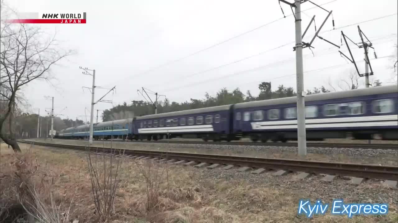 A long train of blue carriages speeds past, its windows reflecting the overcast sky. The NHK World-Japan logo is visible, and the "Kyiv Express" text gives context.
