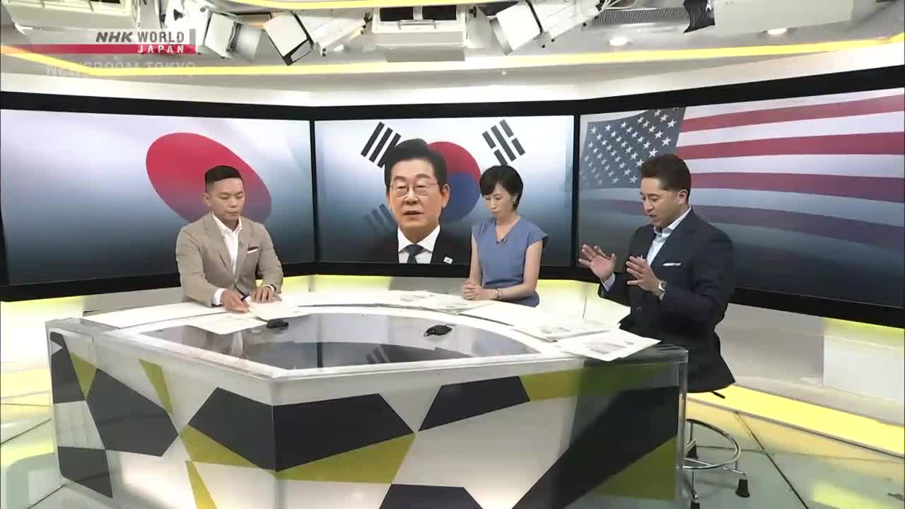 Three people sit behind a geometric desk, with a backdrop of flags and a politician's face on the screens. The man in the suit gestures with his hands as he speaks, likely on NHK World-Japan.
