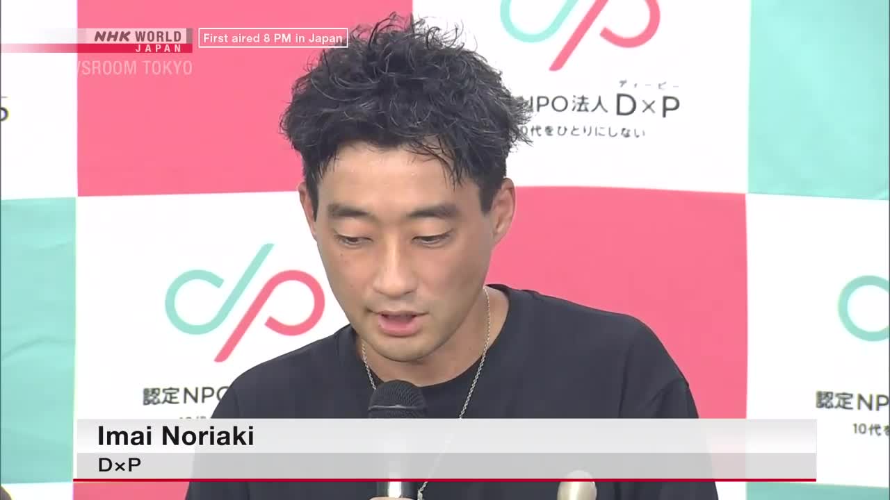 Imai Noriaki, from D×P, speaks into a microphone, his gaze focused downwards. Behind him, the NHK WORLD JAPAN logo and a pink and teal backdrop are visible.
