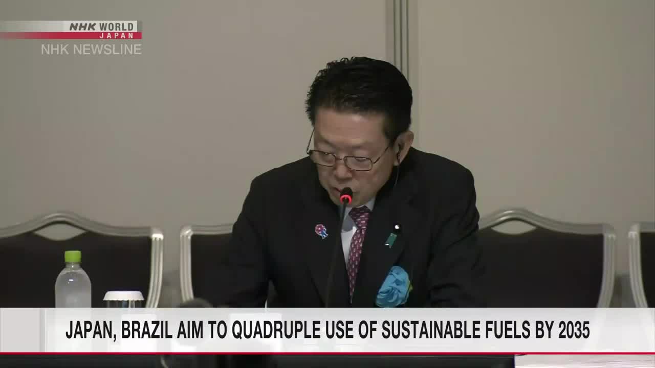 A man in a suit speaks into a microphone, his eyes focused on papers in front of him. The NHK World-Japan news banner displays a headline about sustainable fuels.
