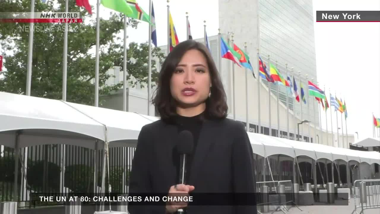 A reporter from NHK World-Japan stands in front of the United Nations building in New York, speaking into a microphone. Behind her, numerous flags of different nations flutter in the breeze.

