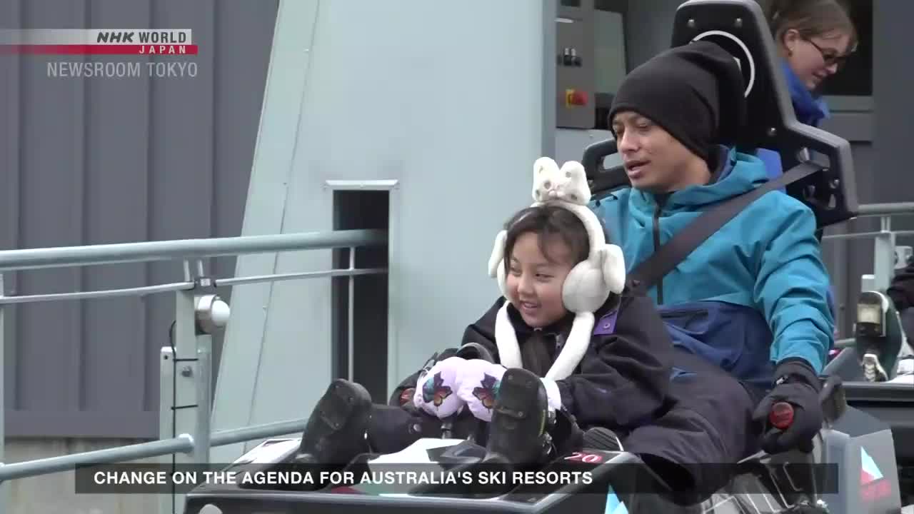A father and daughter, both bundled up, are seated in a ride vehicle, the girl's face lit up with a smile. The NHK World-Japan broadcast, "Newsroom Tokyo," has a graphic about Australian ski resorts at the bottom of the screen.

