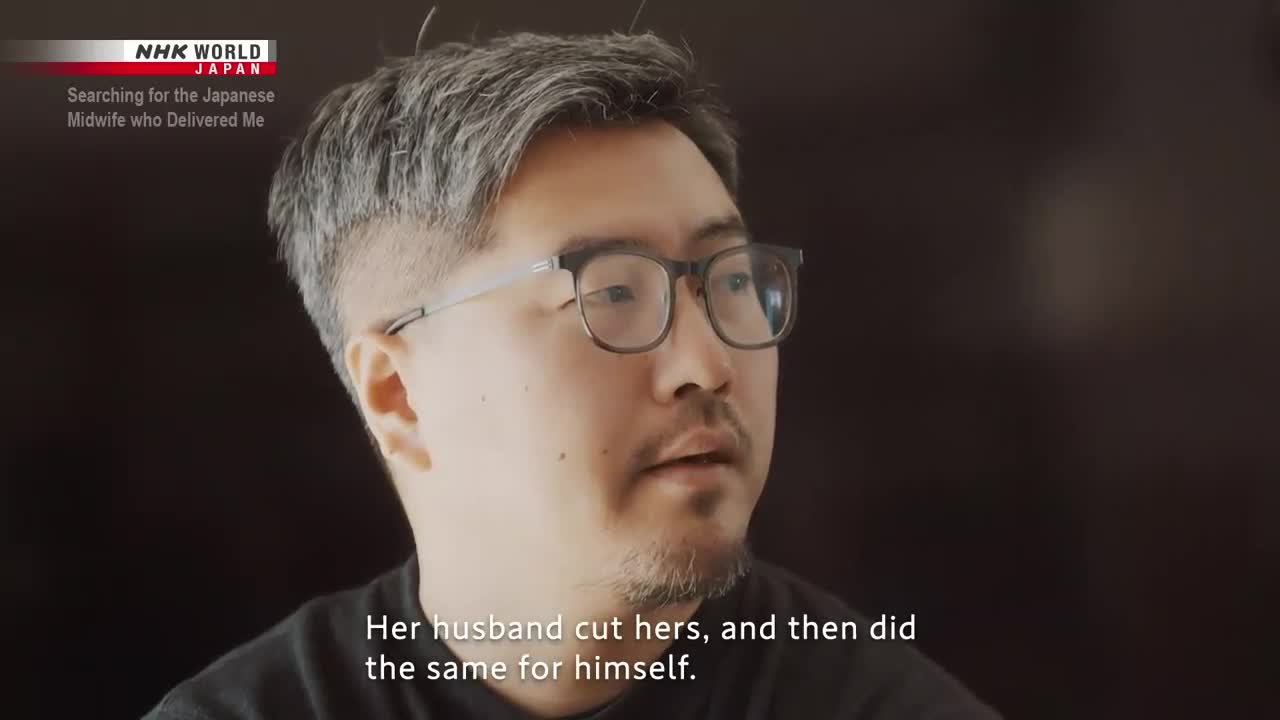 A man, wearing glasses, is speaking, his gaze slightly off-camera. Text on screen, from NHK World-Japan, subtitles a story about a midwife.
