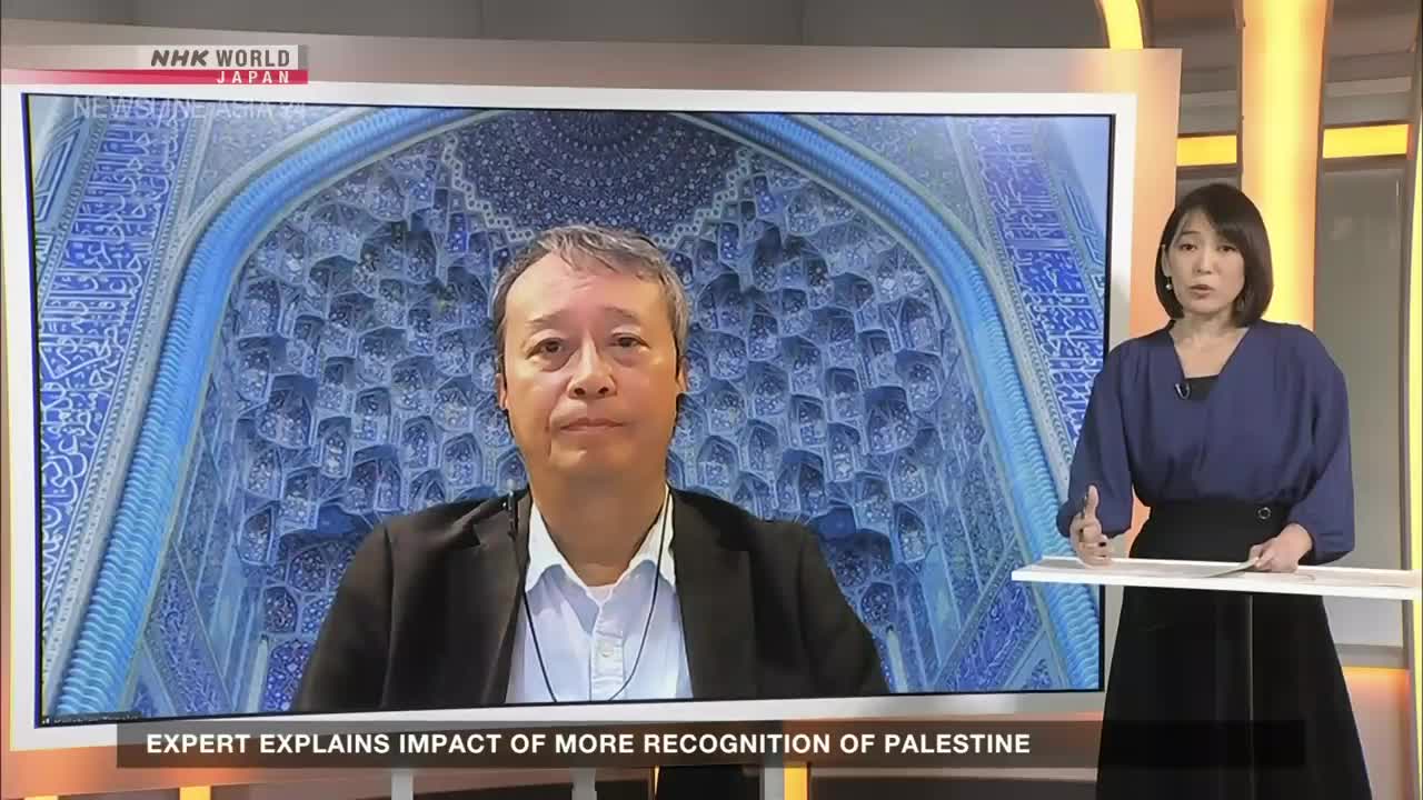 On the NHK World-Japan broadcast, a man in a dark jacket is on screen, speaking in front of a detailed blue and white architectural backdrop. A woman in a blue blouse gestures with her hands as she stands beside the screen.
