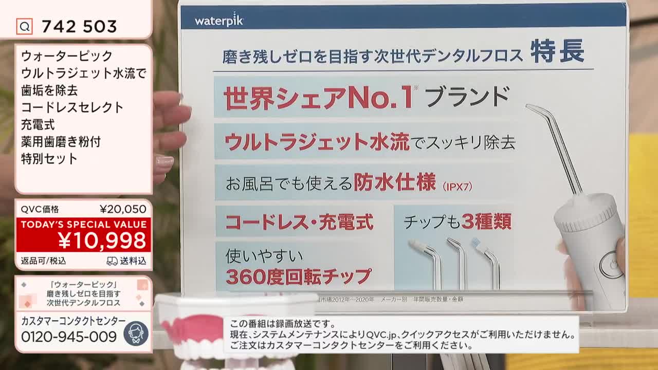 A hand holds a white Waterpik dental flosser, showcasing its features on a Japanese QVC broadcast. The screen displays the product's price, a special offer, and bullet points in Japanese, highlighting its benefits.
