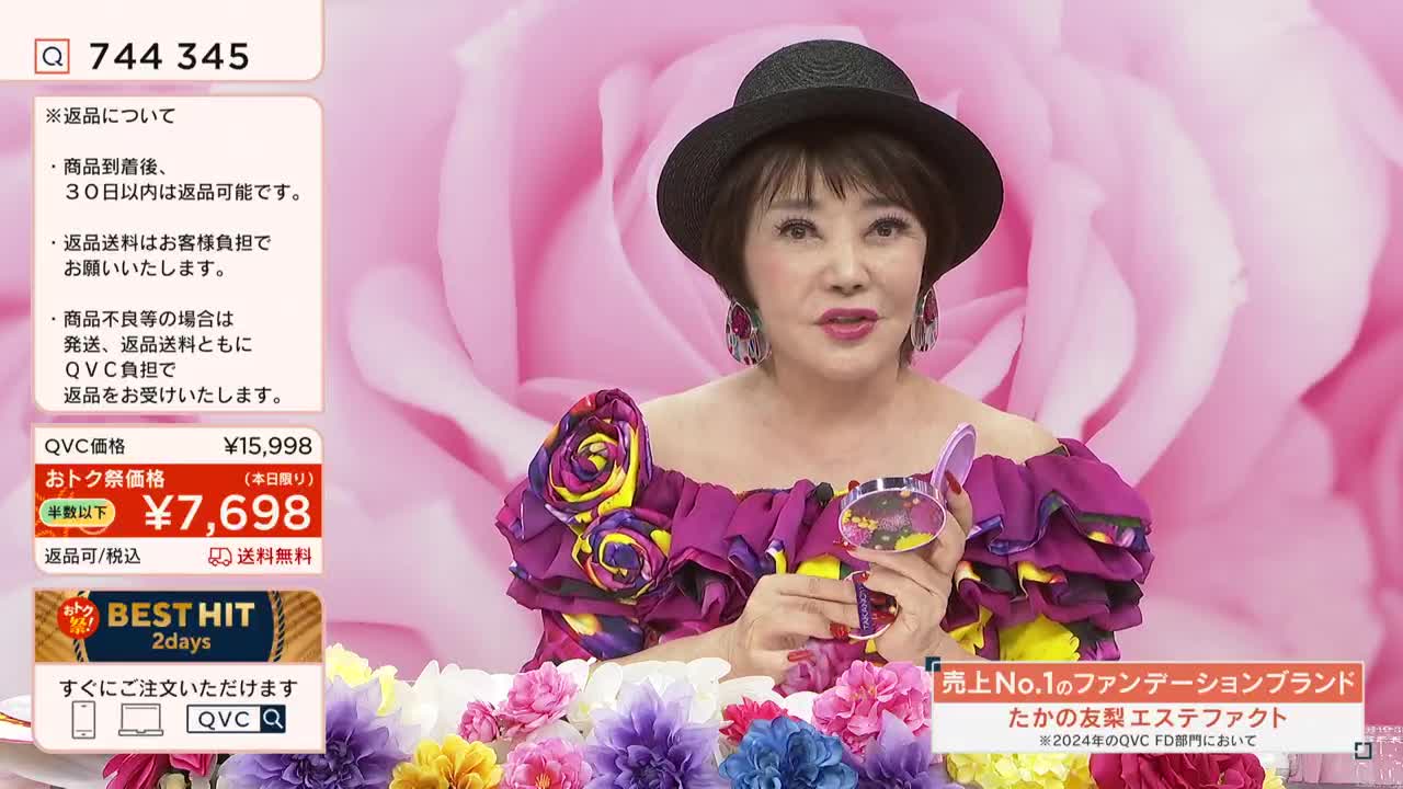 A woman in a hat and a flamboyant top holds a compact, speaking to the camera in a QVC-style broadcast. Beneath her, a floral arrangement adds to the presentation's soft, feminine aesthetic.
