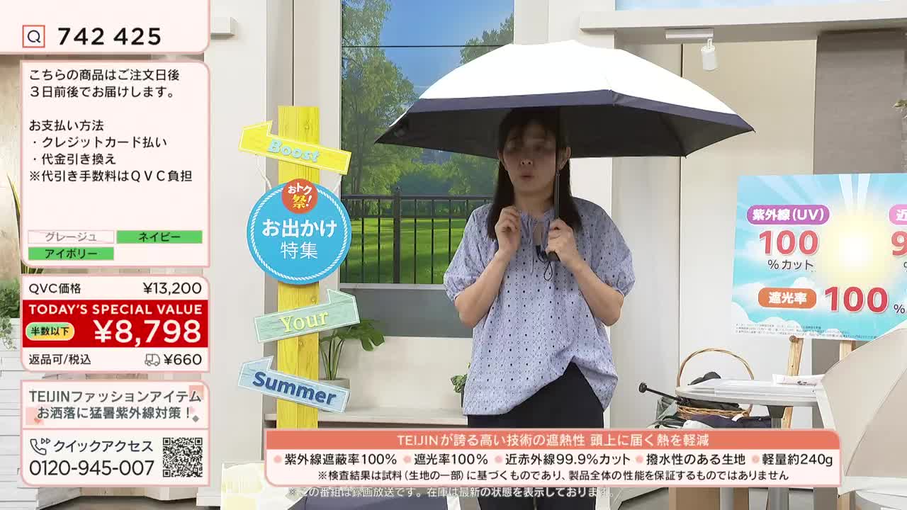 A woman stands in front of a backdrop, holding a partially open umbrella. She's on a QVC-style show, likely in Japan, presenting the product with a serious expression.
