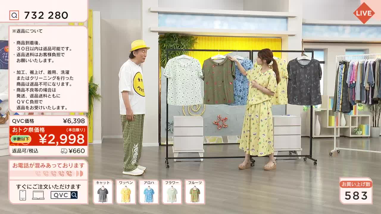 A woman in a yellow dress holds up a shirt, gesturing towards a man in a yellow hat and a smiley-face t-shirt. They're on a QVC-style show in Japan, with a rack of clothes and on-screen pricing information.
