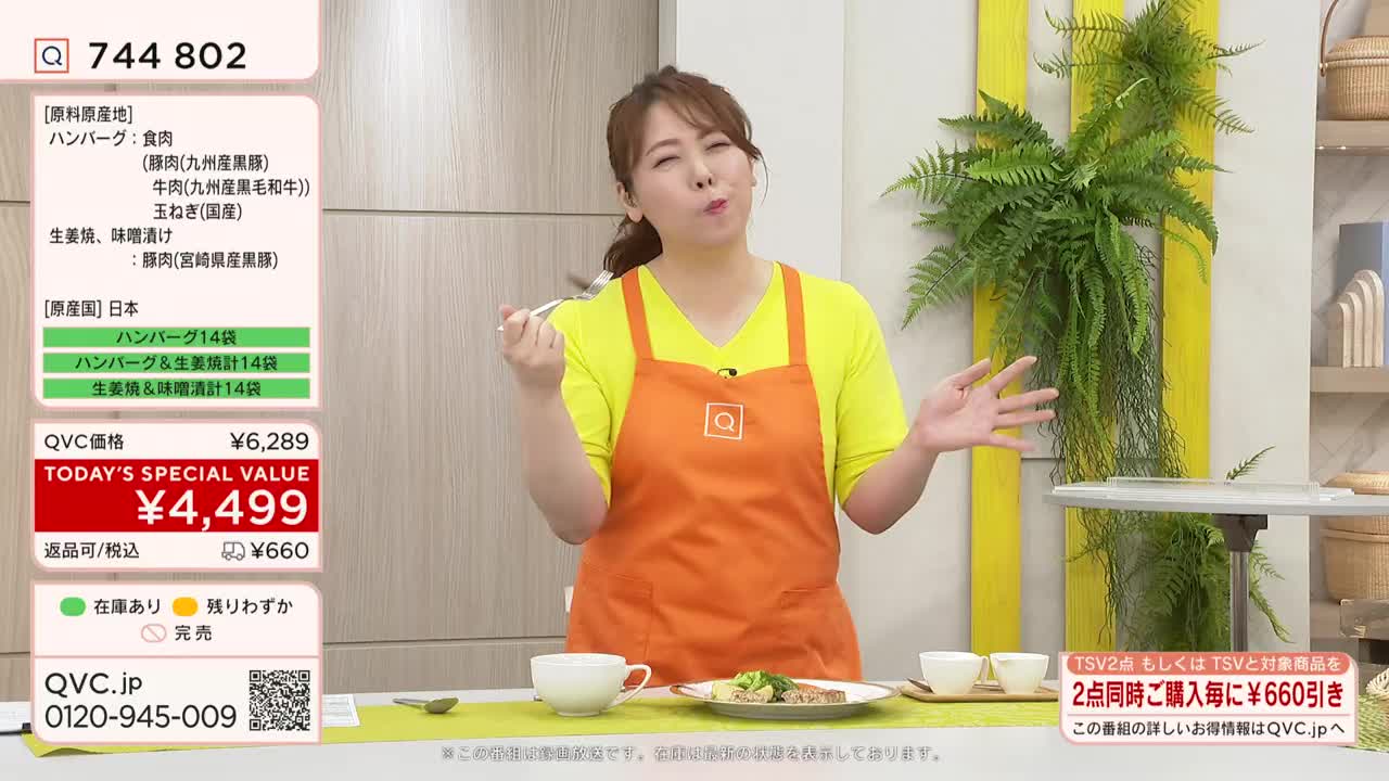 A woman in an orange apron, likely on a QVC Japan broadcast, holds a fork to her mouth. She seems to be tasting something, her expression conveying enjoyment.
