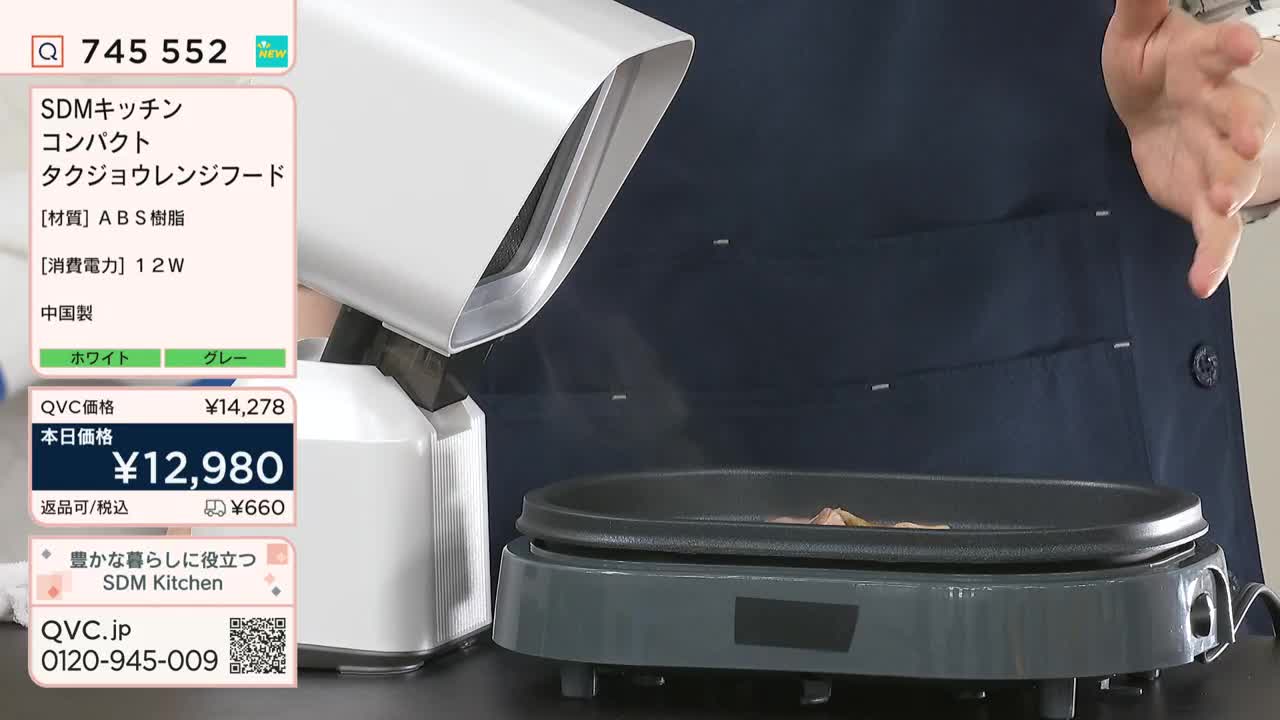 A compact, white kitchen appliance hovers over a dark gray griddle, and steam rises from it.  The presenter gestures towards the cooking surface, likely demonstrating the product on QVC Japan.
