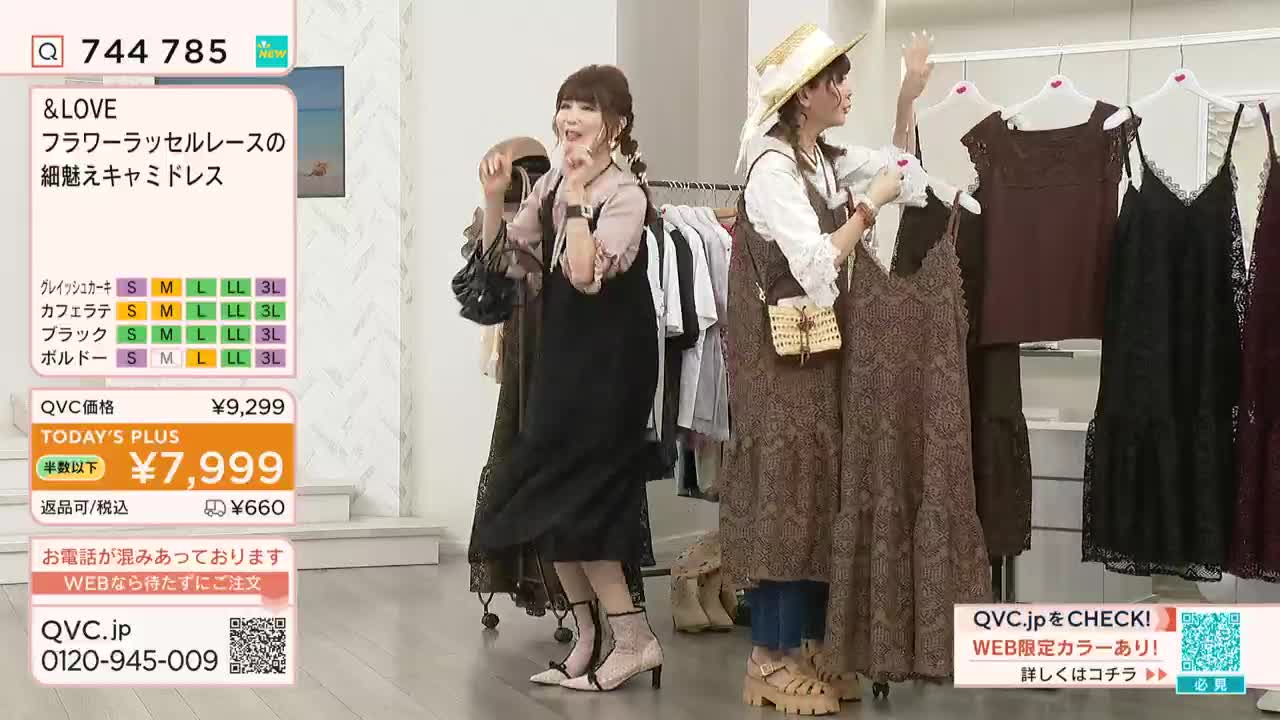 Two women on a QVC set in Japan are gesturing toward the camera, showcasing lace dresses hanging on a rack. The woman on the left is wearing a black dress, and the one on the right is holding up a brown one, both smiling.
