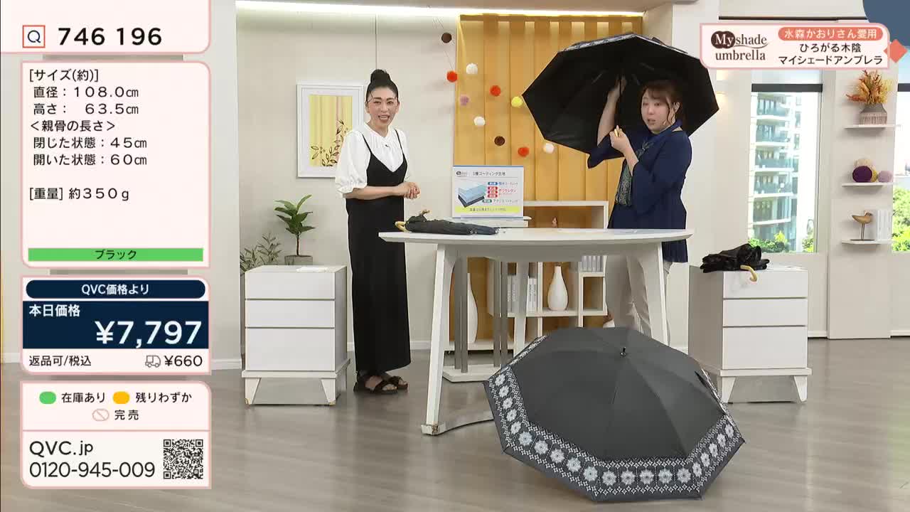 A woman in a navy top holds a black umbrella open, demonstrating its size. Another woman, dressed in black, stands beside a table, ready to provide more details on this QVC Japan product.
