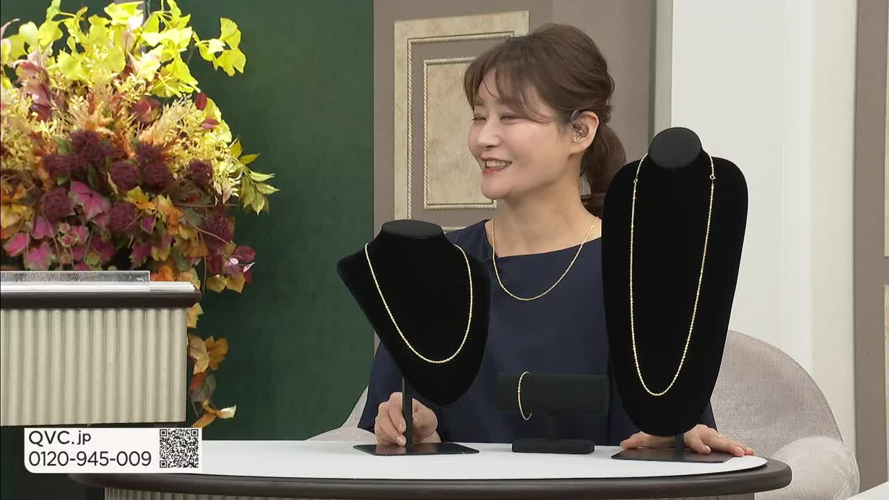A woman on a QVC Japan set smiles, gesturing toward two necklaces displayed on black stands. The camera focuses on the jewelry, with a QR code and phone number visible in the corner.
