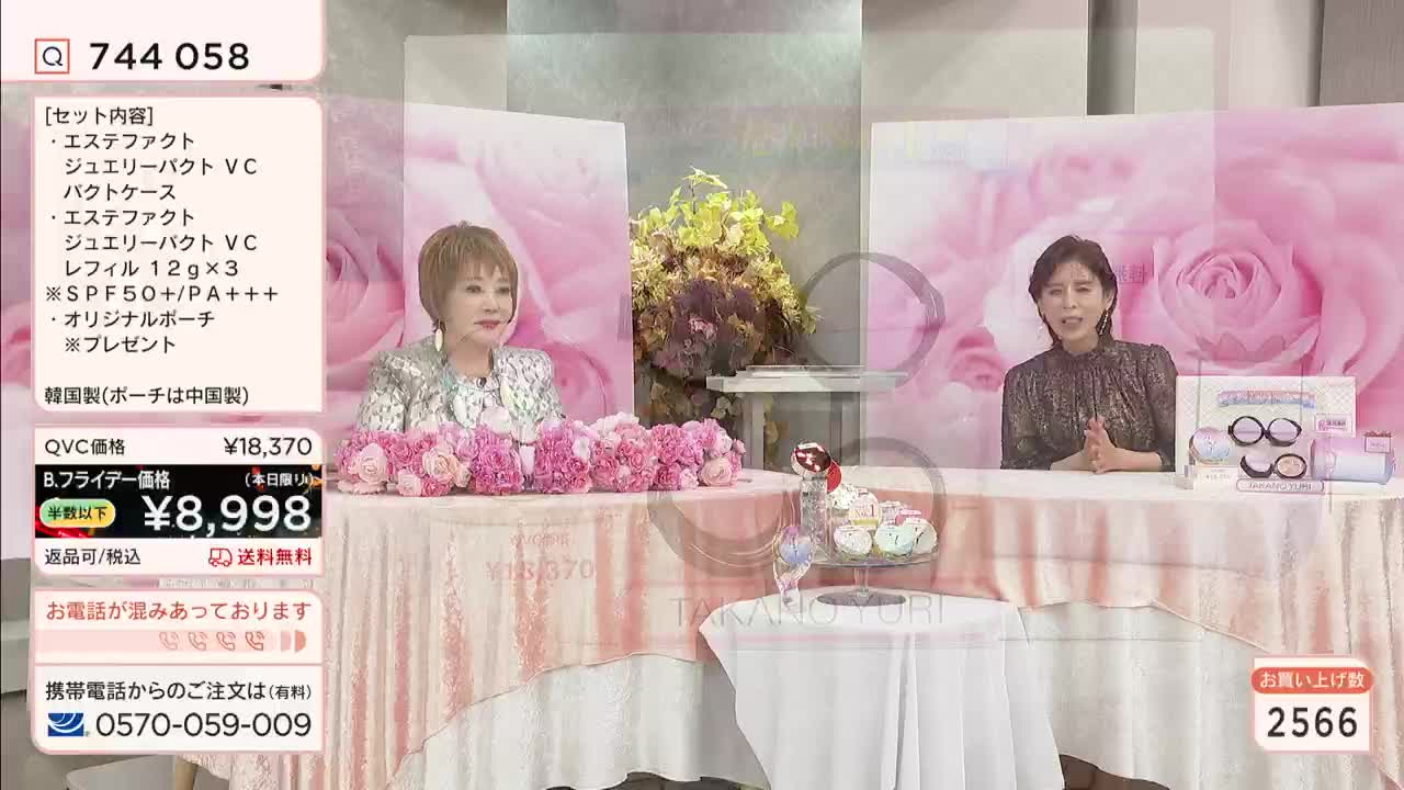 Two women are seated at separate tables, each adorned with a floral arrangement, in a Japanese QVC broadcast. The presenters are discussing a beauty product, with the price and product details displayed on a screen.

