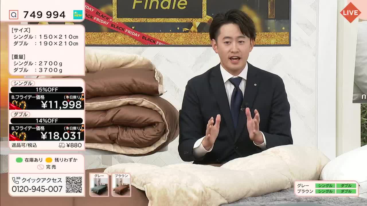 A man in a suit gestures enthusiastically, promoting a brown and beige comforter on a QVC-style show. Beside him, a stack of the comforters sits, and a price list in Japanese is displayed on the screen.
