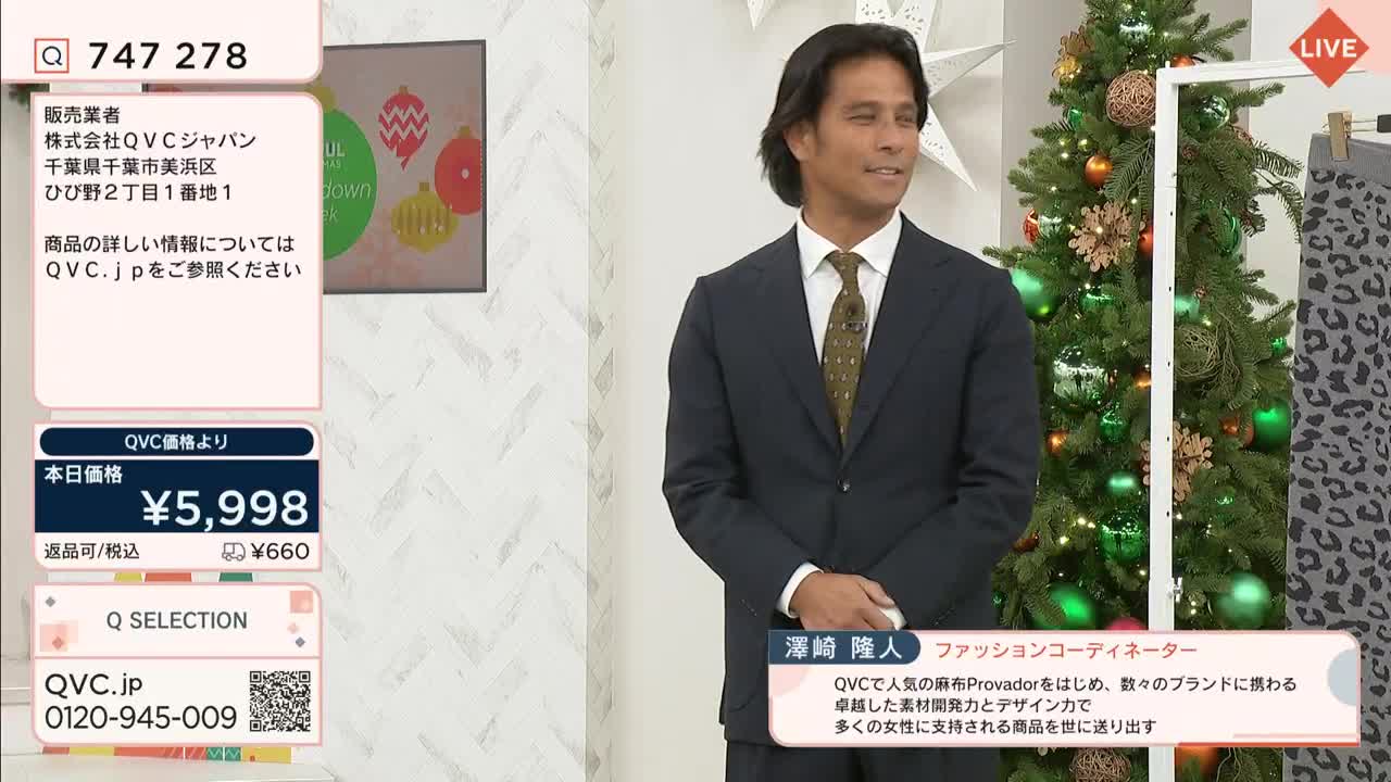 A man in a suit stands in a studio, smiling slightly, with a Christmas tree and a rack of clothes behind him. Graphics from QVC Japan are displayed on the screen to his left, and a price is listed.
