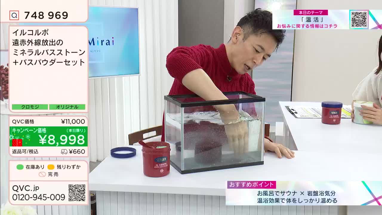 A man in a red sweater is stirring water in a small aquarium on a QVC Japan set. Next to the tank, a jar of bath powder sits, presumably for the demonstration of the product.
