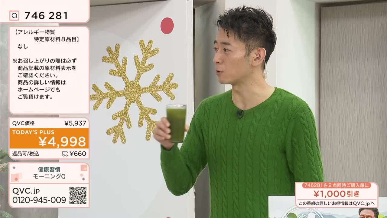 A man in a green sweater holds a glass of green liquid, likely a drink, while speaking on a QVC-style program. Behind him, a large gold glitter snowflake decorates the wall.

