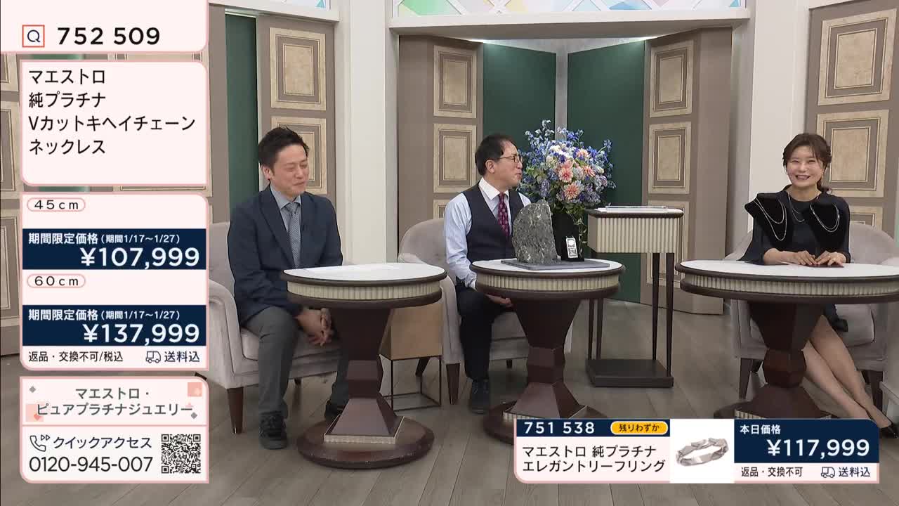 A man in a suit sits to the left, while another man in a vest and tie gestures towards a display item. A woman smiles from her seat on the right, her shoulders adorned with a striking black collar.