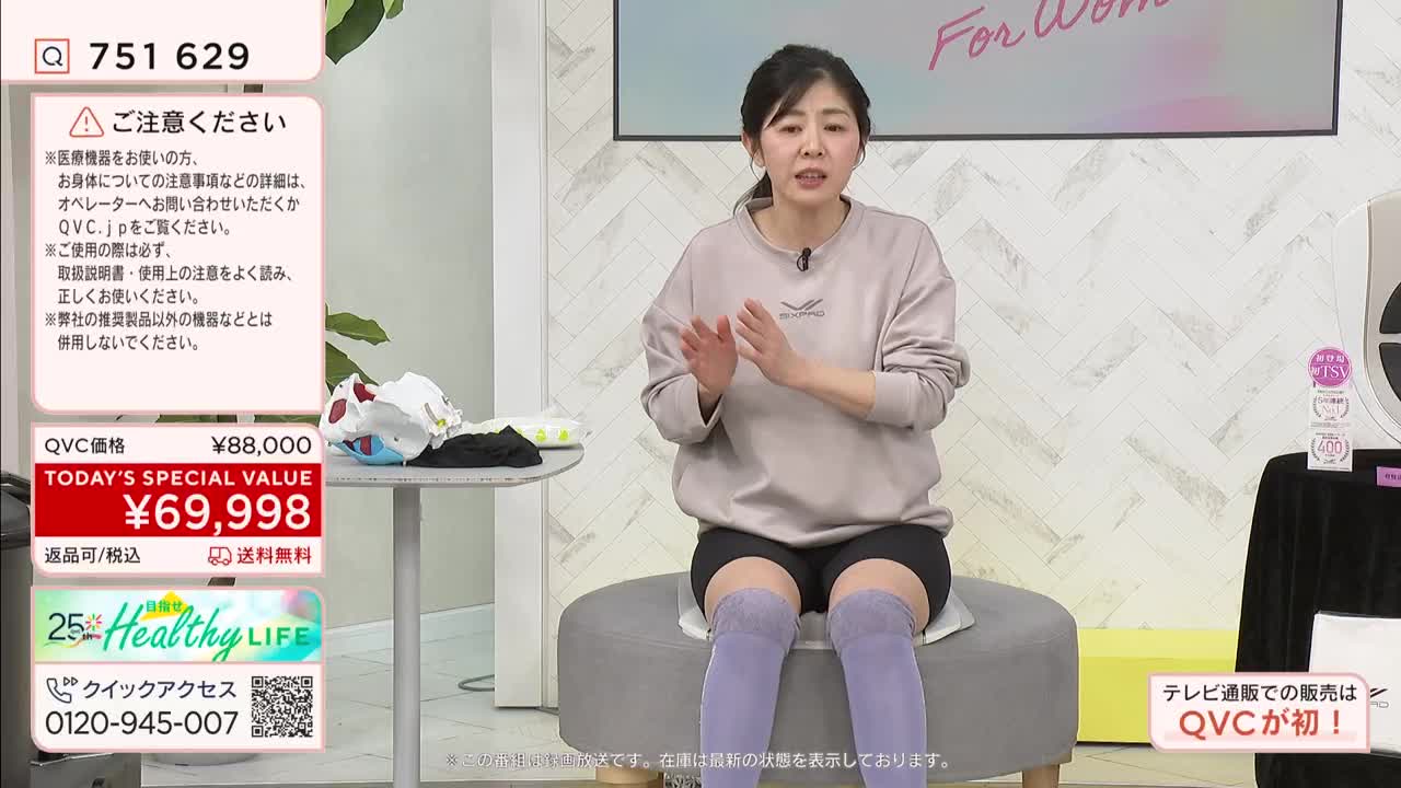 A woman on a QVC broadcast in Japan gestures with her hands while sitting. On the table next to her, a pile of white and black fabric items rests. A woman on a QVC broadcast in Japan gestures with her hands while sitting. On the table next to her, a pile of white and black fabric items rests.