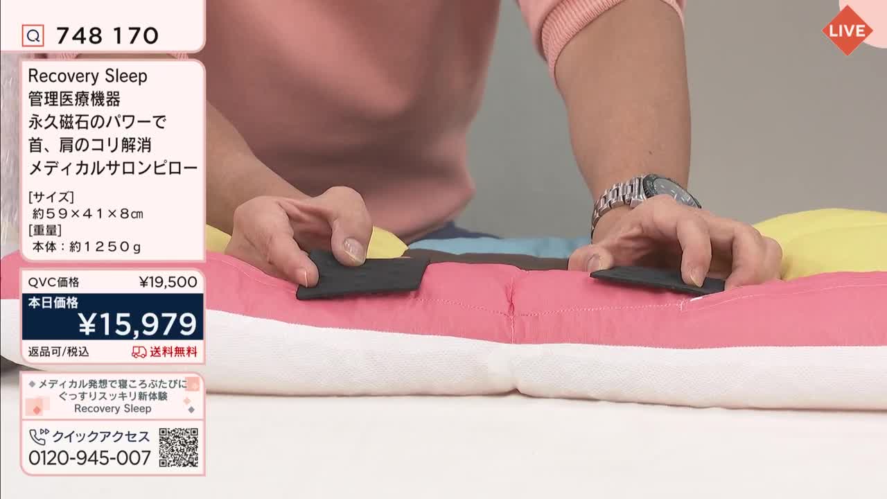 Someone is demonstrating a recovery pillow on QVC Japan. They press down on two black rectangular pads on the pillow's surface.