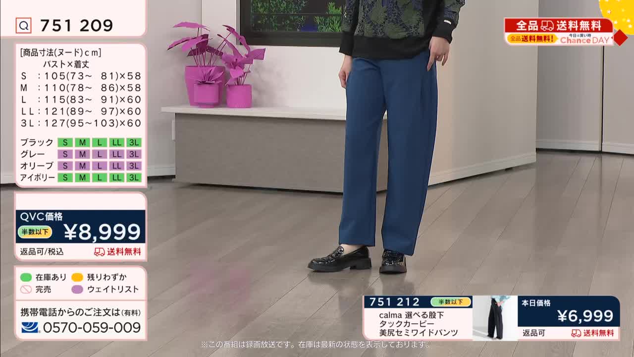 A woman stands in a studio, her dark blue pants pooling slightly over black loafers. The QVC broadcast displays product details and pricing for the garment.