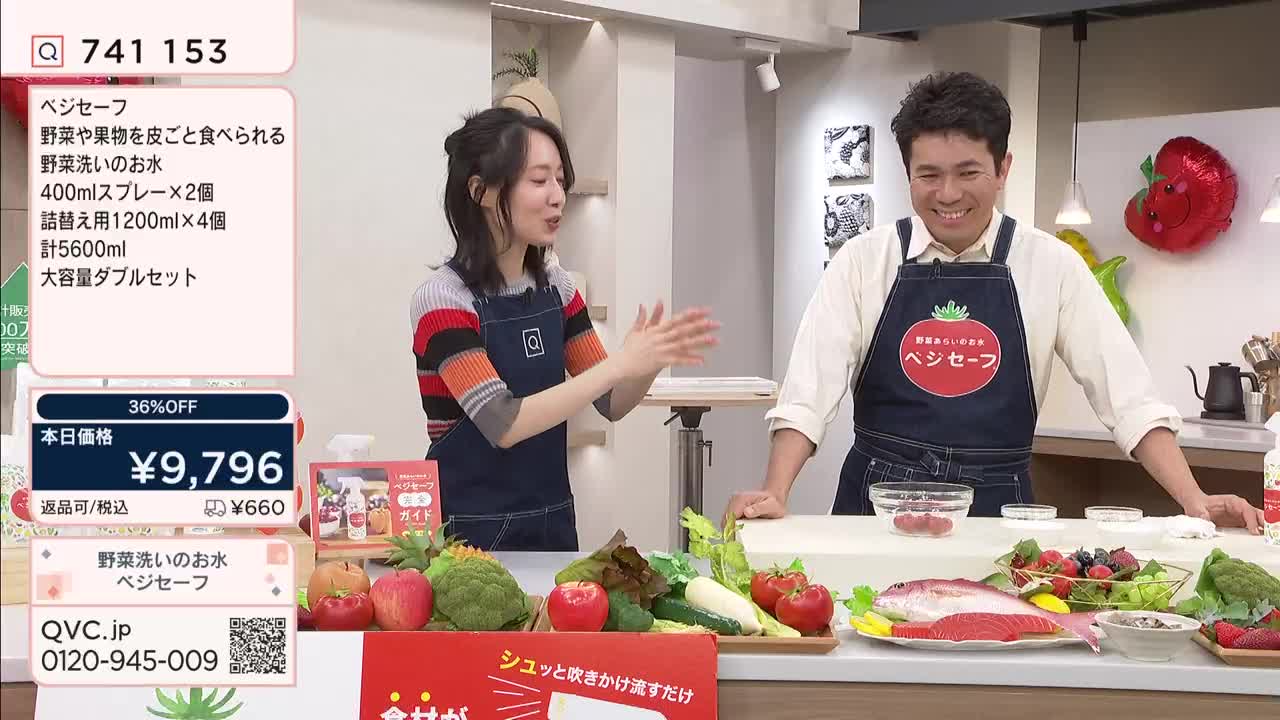 A woman claps her hands together while a man smiles, both wearing aprons. A display of fresh produce and a whole fish sits on the counter between them.