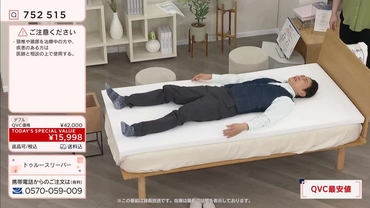 A man lies on his back on a white mattress topper. He's wearing a blue vest over a white shirt and dark pants, his arms and legs spread wide. A graphic on the left shows the item's price, ¥15,998, as a "TODAY'S SPECIAL VALUE" from QVC Japan.