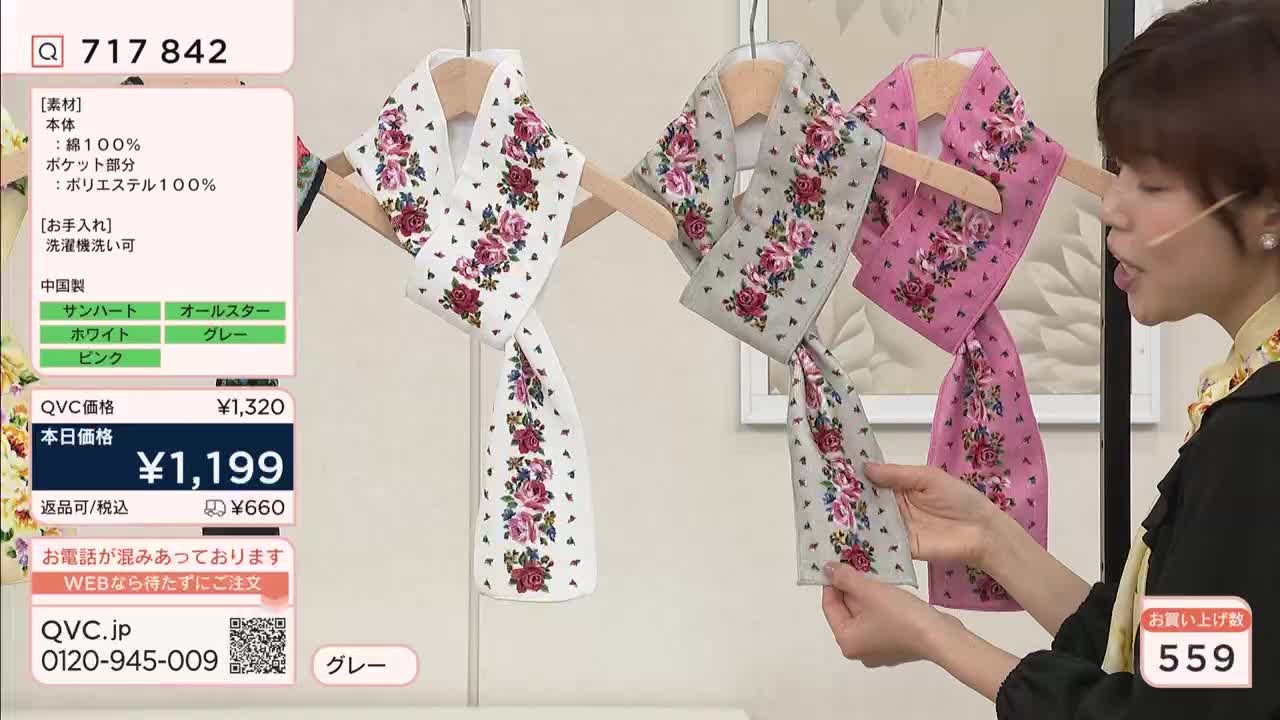A woman on QVC Japan holds up a grey scarf with a floral pattern. Three scarves, white, grey, and pink, hang on display, each adorned with a band of roses.