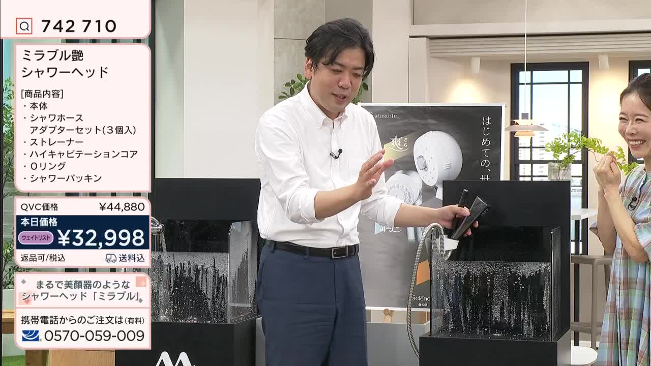 A man in a white shirt gestures with a showerhead, demonstrating its features over a display tank. A woman beside him smiles, holding a plant.
