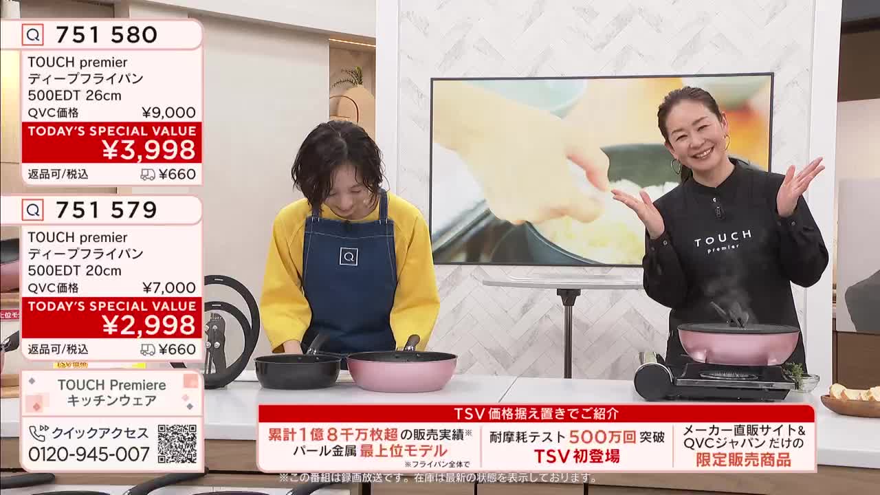 A woman in a yellow shirt and blue apron bends over two pans, one black and one pink, on a white counter. Beside her, another woman in a black shirt smiles with her hands outstretched as steam rises from a pink pan on a portable burner.