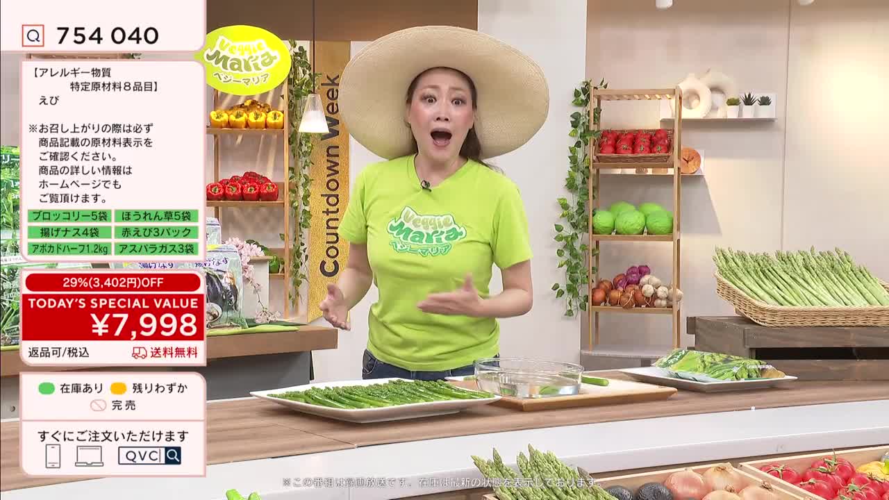 A woman in a bright green shirt and a wide-brimmed hat gestures emphatically while presenting a platter of asparagus. Behind her, shelves display colorful produce, and a basket overflows with more fresh green stalks.