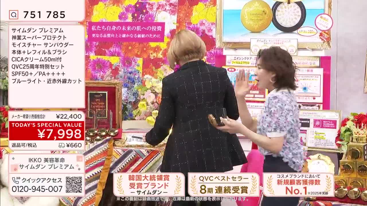 A woman in a patterned blouse gestures enthusiastically while facing away from the camera. Behind her, a woman in a dark jacket holds a small gold object.