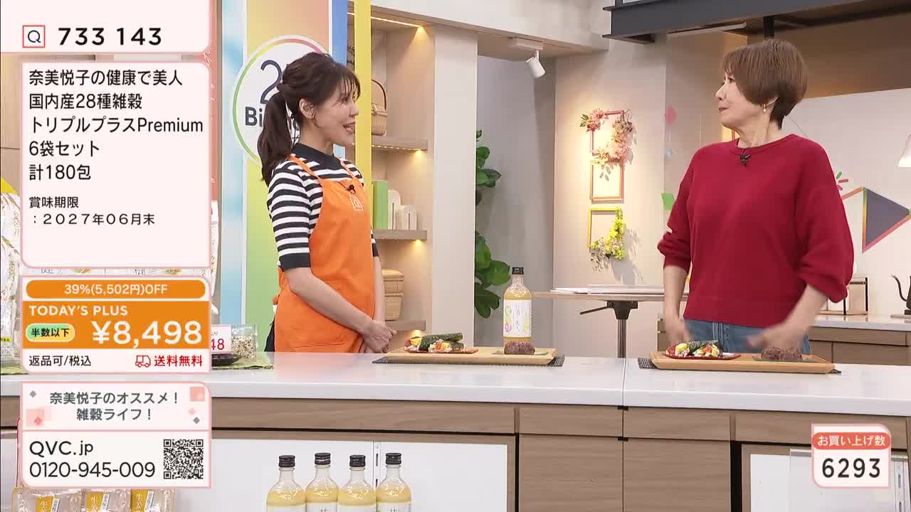 A woman in an orange apron stands beside a counter, gesturing towards a display of food. Another woman in a red sweater looks on, her hands clasped in front of her.