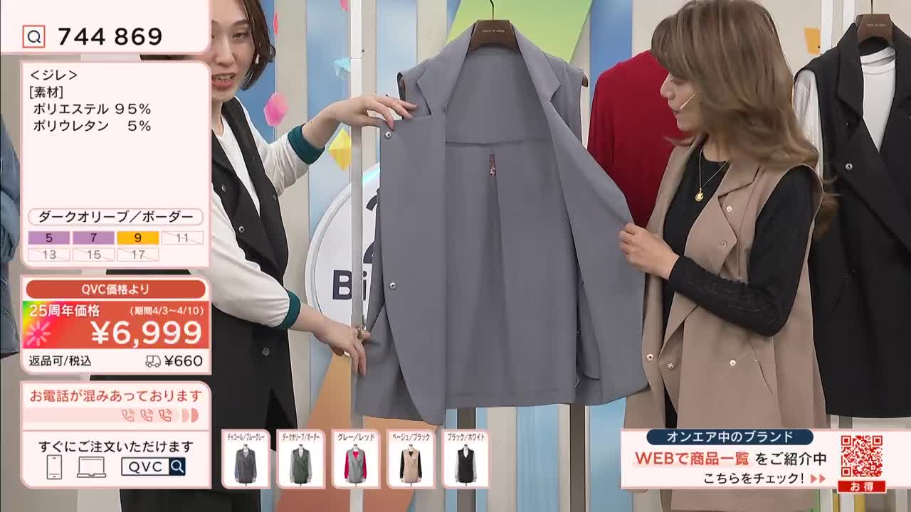 Two women on a QVC broadcast in Japan are presenting a sleeveless grey jacket. One woman holds the jacket open, showing its lining, while the other gestures towards it.