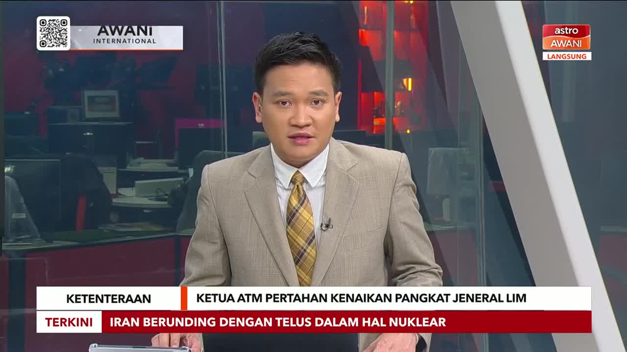 A man in a tan suit and patterned tie is speaking directly to the camera. The Astro Awani logo is visible in the corner, and headlines scroll across the bottom of the screen.
