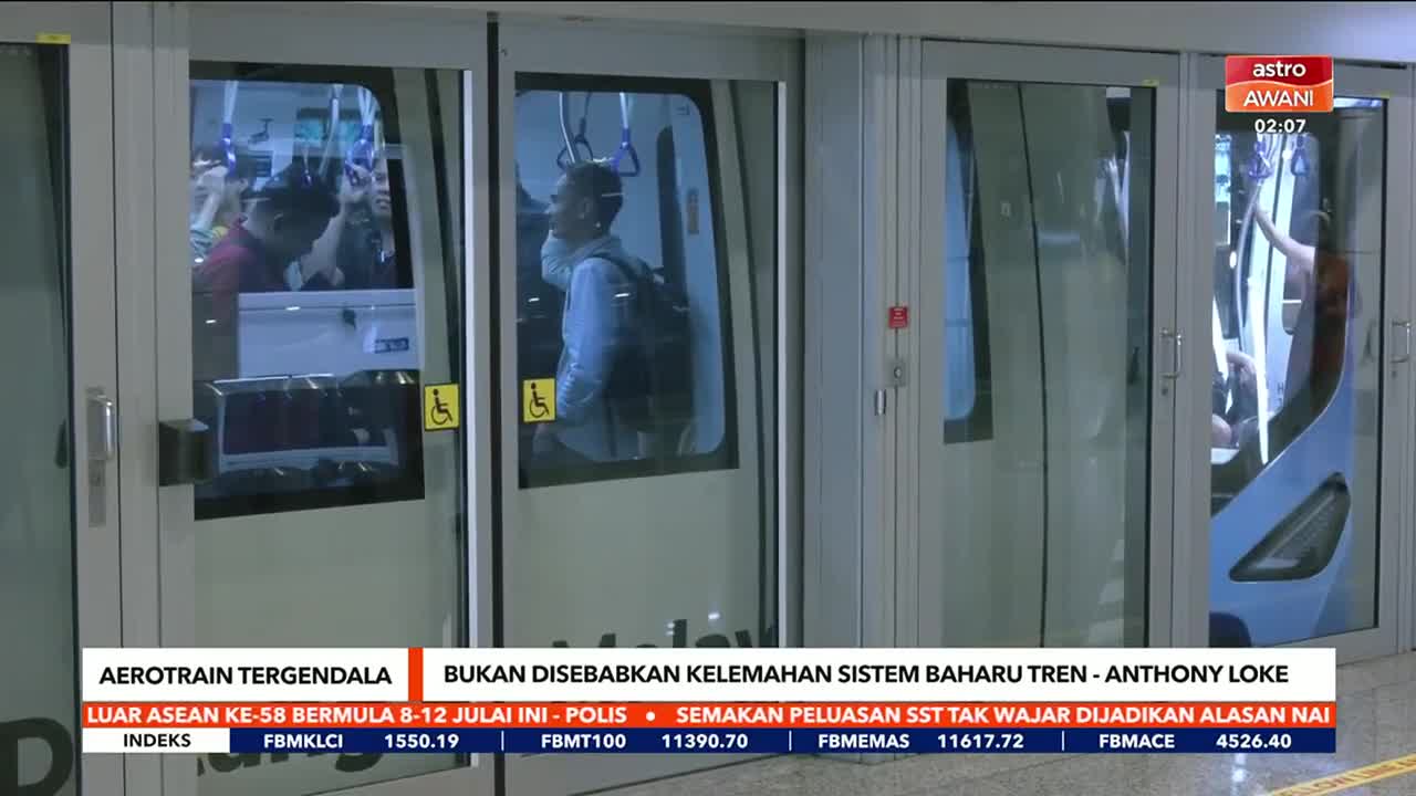 Astro Awani is reporting on a stalled aerotrain; passengers are visible inside the train's doors. A man with a backpack stands near the glass, and the doors are closed.
