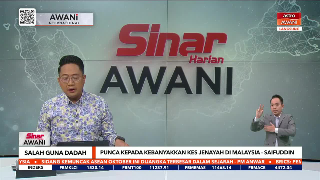 A man in a patterned shirt speaks at a desk, while another man, interpreting, uses hand gestures on the right. The Astro Awani logo and a map of Malaysia are visible in the background.
