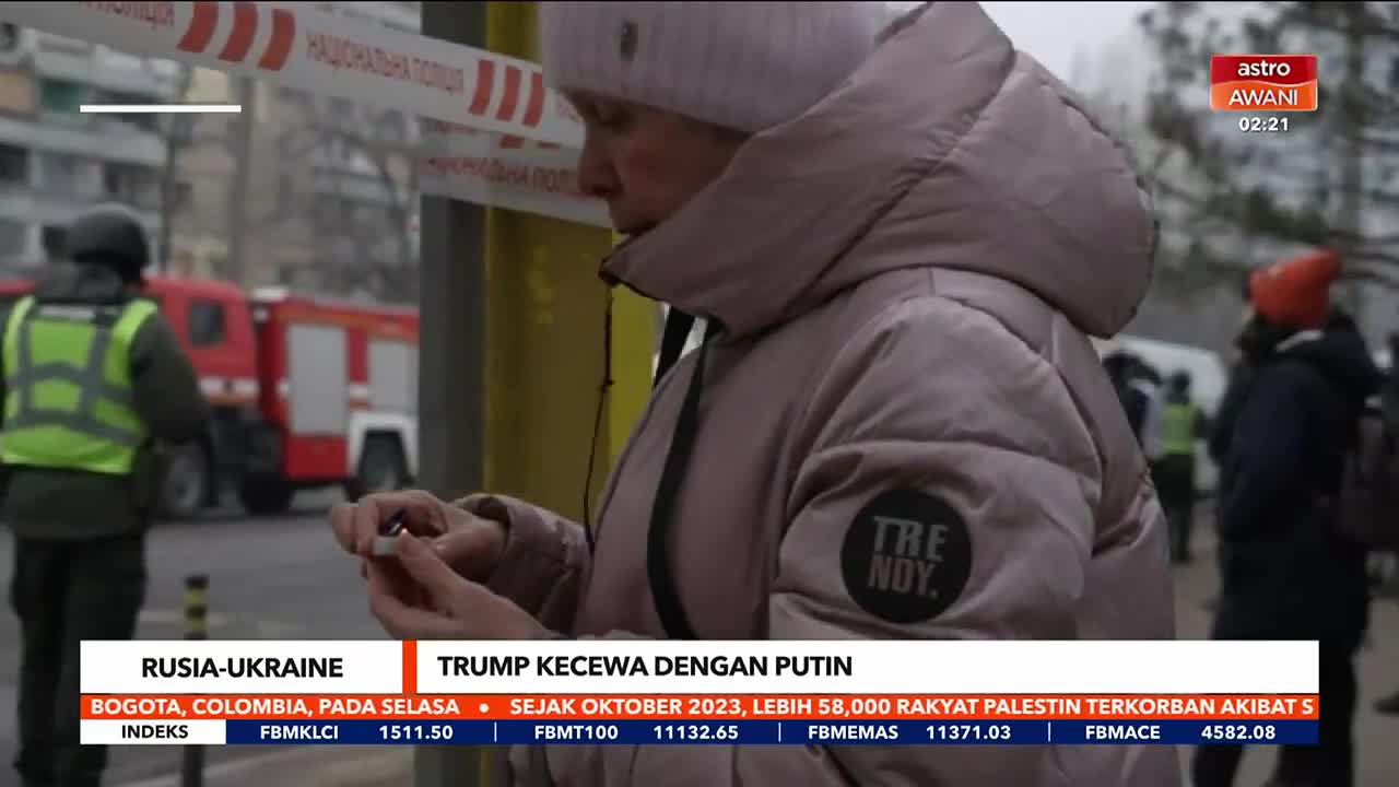 A woman in a puffy coat and a beanie fiddles with something small in her hands, her gaze fixed downward. Behind her, a fire truck sits near a police barricade, the scene likely captured for Astro Awani.
