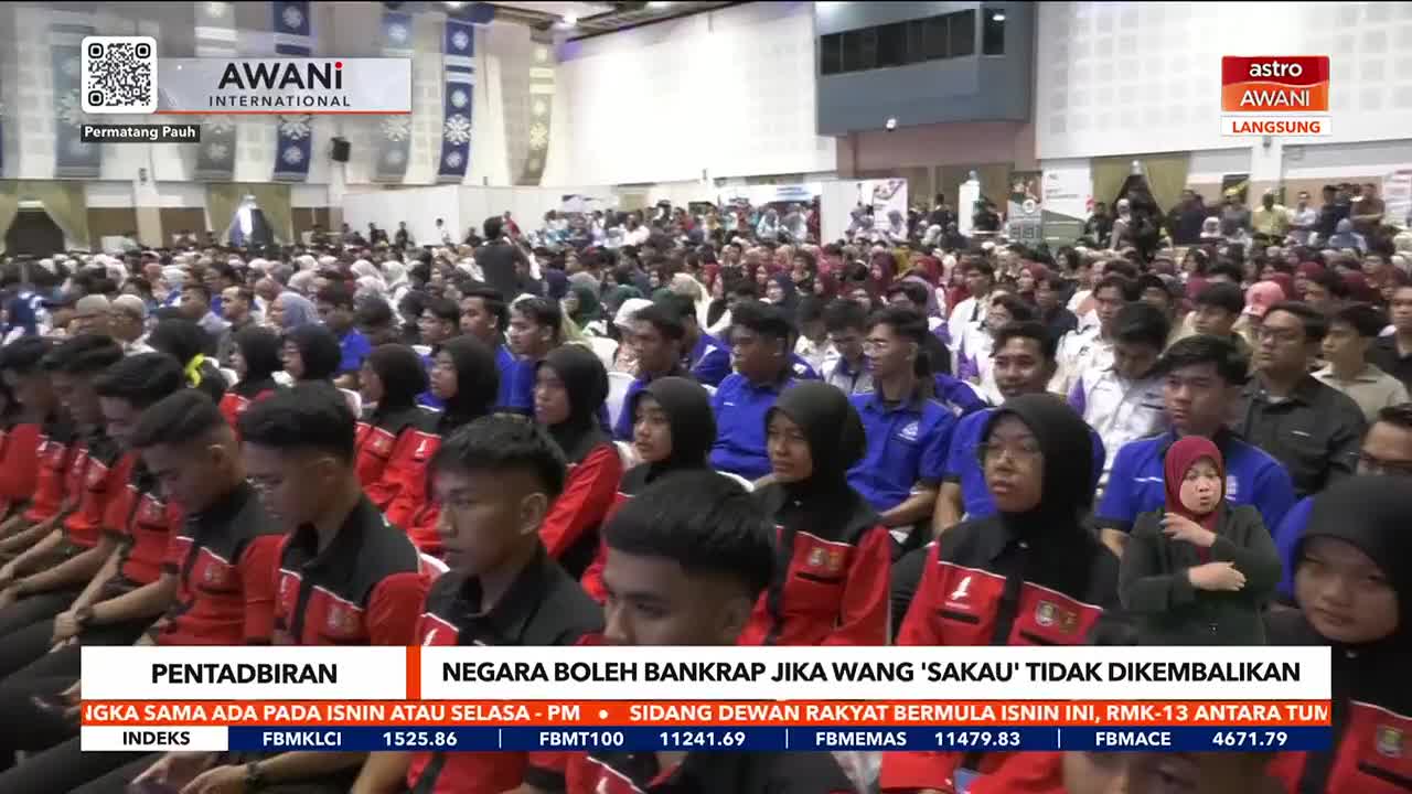 A large crowd sits in a hall, many wearing red and black jackets, while a live broadcast from Astro Awani plays on a screen. The news coverage is reporting on a discussion about Malaysia's financial state.
