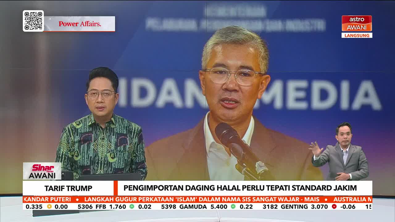 A man in a suit speaks into a microphone, his image large on the screen, while two other presenters flank him on either side. The Astro Awani logo is visible in the corner, and the news ticker scrolls across the bottom.
