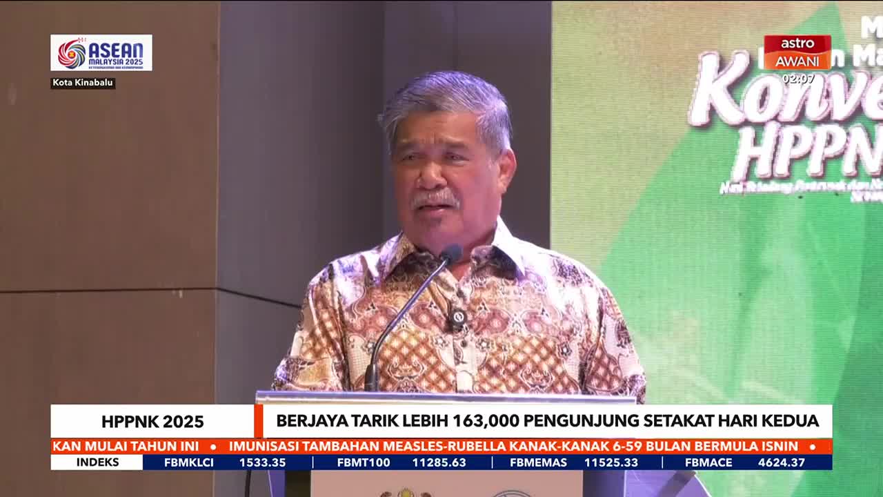 A man in a patterned shirt speaks at a podium, the Astro Awani logo visible above his head. Below, a ticker displays financial information and the headline "BERJAYA TARIK LEBIH 163,000 PENGUNJUNG."
