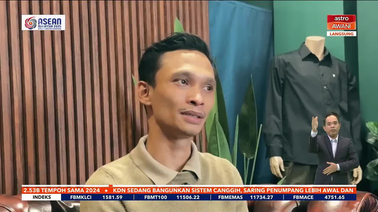A man in a tan shirt is speaking on Astro Awani, with the ASEAN Malaysia 2025 logo visible in the background. Below, the financial index tickers scroll across the screen.
