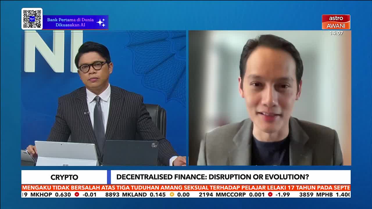 On Astro Awani, a man in a suit and tie sits at a desk, while another man appears on a screen, likely in a video call. The broadcast is discussing crypto and decentralized finance.
